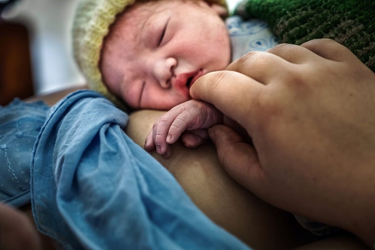 A newborn baby lays on her mother. The mother holds the baby's hand.